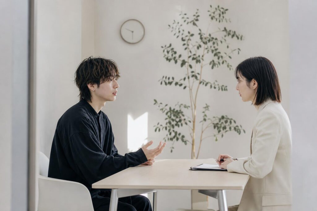 小さな面接ブースで、応募者役の美容師がテーブル越しに採用担当と向かい合い、少し肩をすくめながら言葉を選んでいる。カメラは二人の横顔が見える位置からミドルショットで捉え、背景には観葉植物と壁掛けの時計がぼんやり写り込み、緊張感のある静けさを強調している。