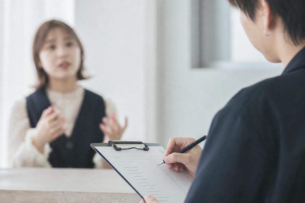 面接室の隅、二人が対面で座り、面接官役の人物は右手に持ったペンで手元のチェックリストに小さく丸をつけながら、正面に座る求職者の身振り交じりの説明に耳を傾けている。カメラは真横からのクローズアップショットで、面接官の指先とチェックリストの余白にピントを合わせ、背景の求職者の上半身は柔らかくぼやけて写り込み、評価する側の視点が強調される構図になっている。
