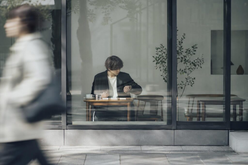 サロン近くの喫茶店の窓際の一人用テーブルで、開始前の一人の人物が小さなカード三枚に「技術方針」「客層」「教育体制」と書き分け、テーブルの端に置いた腕時計にちらりと視線を送っている。カメラは店外の歩道側からガラス越しに捉えたロングショットで、通行人の影が手前にぼんやりと写り込み、面接直前の静かな緊張感を距離感のある構図で表現している。