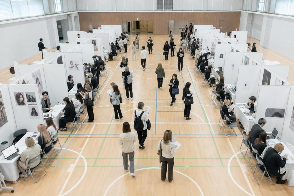 体育館を利用した合同説明会の会場全体を見渡すショットで、等間隔に並んだ十数のブースの前に数人ずつの学生が集まり、それぞれ異なるサロンの担当者から説明を受けている。カメラは会場後方の高い位置からの俯瞰広角ショットで、手前のブースの列と奥のブースの列を同一フレームに収め、限られた時間で複数のブースを移動しようとする学生たちの動線が浮き上がる構図になっている。