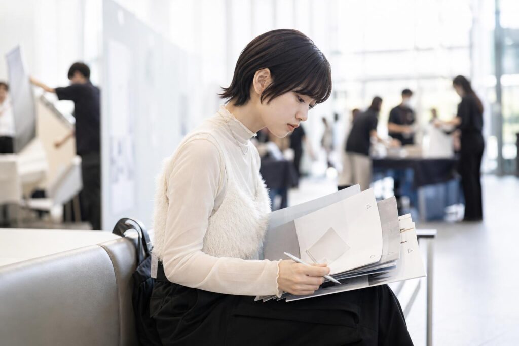 合同説明会が終わった直後の会場出口付近のベンチで、一人の学生が配布されたパンフレット数冊を膝の上に広げ、表紙の余白にペンで小さく丸や三角の印をつけながら見比べている。カメラは正面やや低い位置からのミドルショットで、パンフレットの印と学生の真剣な横顔にピントを合わせ、背景では撤収を始めるブースの様子がぼんやりと写り込み、次の行動を選び取る静かな判断の瞬間を描く構図になっている。
