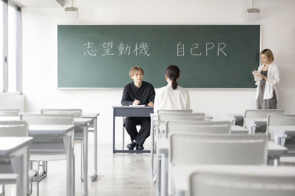 美容専門学校の空き教室で行われている模擬面接の場面。机を挟んで面接官役の生徒が一人、その正面に求職者役の生徒が一人座り、少し離れた壁際で観察役の生徒が一人メモを取りながら立って見ている。教室前方の黒板には「志望動機」「自己PR」という二つの文字が大きく書かれている。カメラは教室の最後列からの引き気味のロングショットで、三人の位置関係と黒板の文字を同一フレームに収め、実践形式で問いの意図を学び合う教室全体の雰囲気を伝える構図になっている。