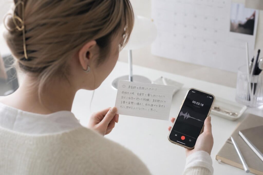 自宅の机に向かった一人の人物が、スマートフォンのボイスメモを起動し、手元の小さなメモカードに書かれた出来事を声に出して話している。カメラは背後斜めから人物の肩越しに捉えたオーバーショルダーショットで、スマホの録音画面と手元のカードにピントを合わせ、背景の壁に貼られたカレンダーはぼんやりとぼやけ、暗記ではなく自分の言葉を確かめるように話す静かな練習の様子を強調する構図になっている。