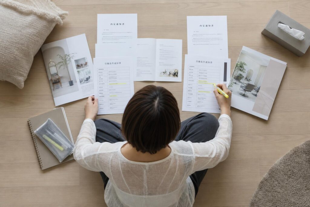 自宅のリビングの床に座り込んだ一人の人物が、二社分の内定通知書とパンフレットを左右に並べ、蛍光ペンで気になる条件に線を引きながら見比べている。カメラは真上からのトップダウンショットで、床に広げられた書類全体と人物の腕の動きを一枚に収め、背景には座布団やティッシュ箱がぼんやりと写り込み、複数の情報を俯瞰的に整理する静かな検討の時間を強調する構図になっている。