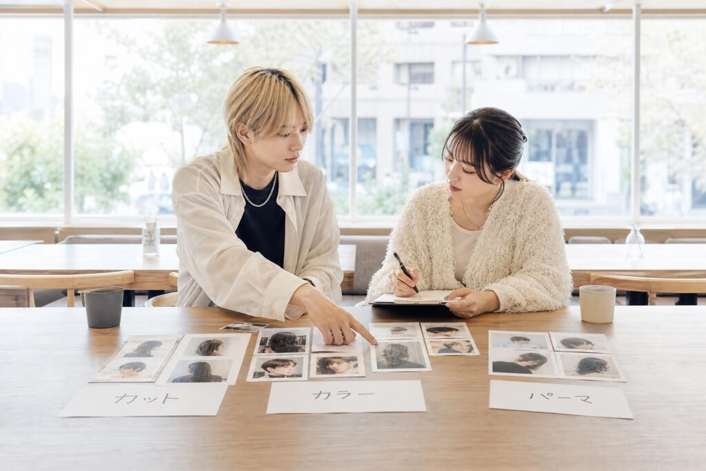 喫茶店の広めのテーブルを挟んで向かい合った二人の人物が、印刷された作品写真の束を「カット」「カラー」「パーマ」と書かれた紙の上にそれぞれ仕分けながら並べ、一人が写真を指差し、もう一人がその組み合わせをメモしている。カメラは正面やや高い位置からのミドルショットで、テーブル全体に広がる写真の配置とメモ用紙を同一フレームに収め、背景の窓外の街並みは柔らかくぼやけて写り込み、技術の幅を意識して構成を組み立てていく検討の過程を際立たせる構図になっている。