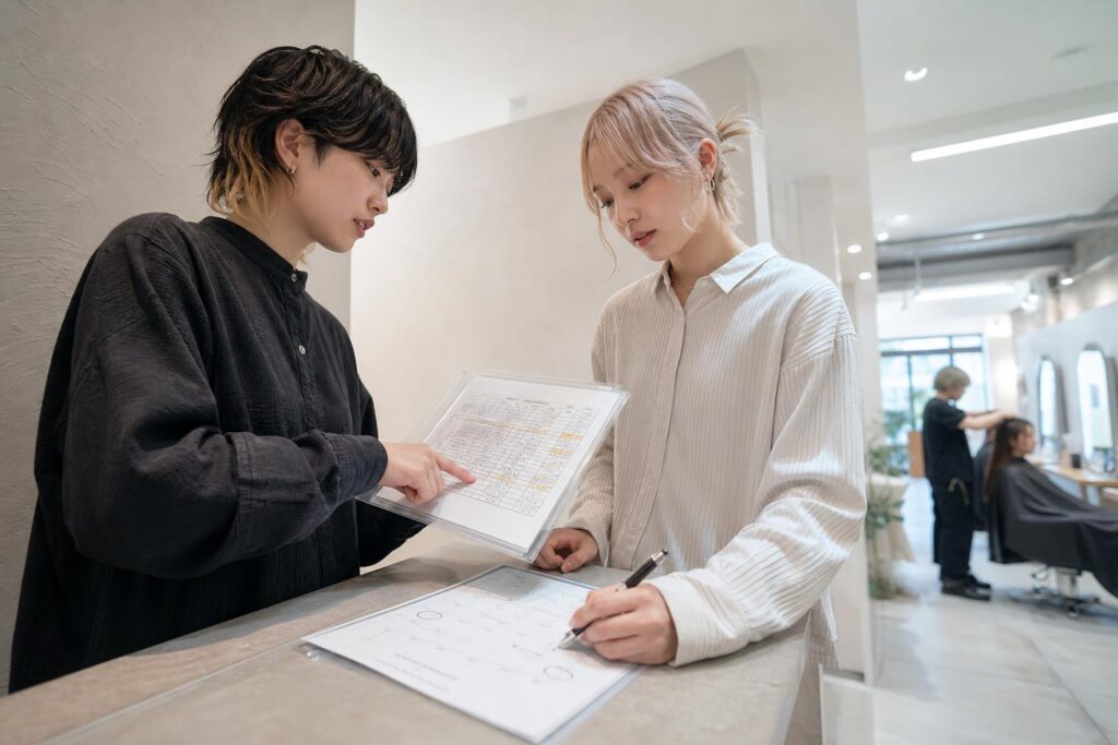 サロンの受付カウンター横で、向かい合って立つ二人の人物のうち一人がクリアファイルに挟んだ顧客リストの表を指でなぞり、もう一人がスケジュール表に稼働日の候補を丸で囲んでいる。カメラは受付側からの斜め下からのローアングルで、二人の腕の動きとファイル・スケジュール表の紙面を同一フレームに収め、背景では奥のセット面でスタッフが施術を続ける様子が縦の奥行きの中にぼんやりと写り込み、引き継ぎ条件と稼働の自由度を同時に具体的に詰めていく実務的なやり取りの緊張感を強調する構図になっている。