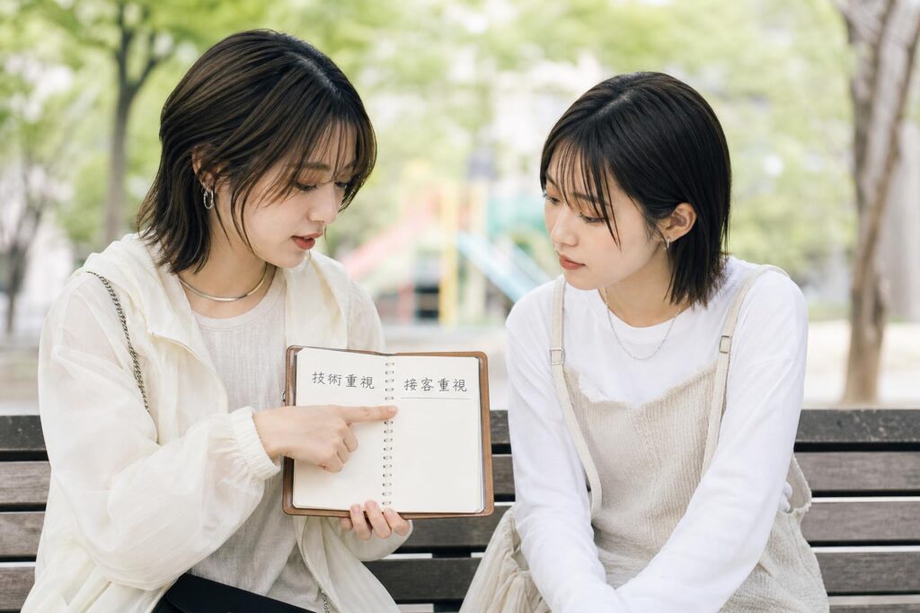 サロン見学を控えた二人の人物が、公園のベンチに並んで腰掛け、一人が手帳に「技術重視」「接客重視」と書いた二つの欄を指差しながら話し、もう一人がその欄を見ながら小さくうなずいている。カメラは正面からの近距離のクローズアップショットで、二人の横顔と手帳の書き込みを同一フレームに収め、背景の木々や遊具はやわらかくぼやけて写り込み、焦って決めるのではなく自分の希望を言葉にして確かめ合う落ち着いた対話の様子を強調する構図になっている。