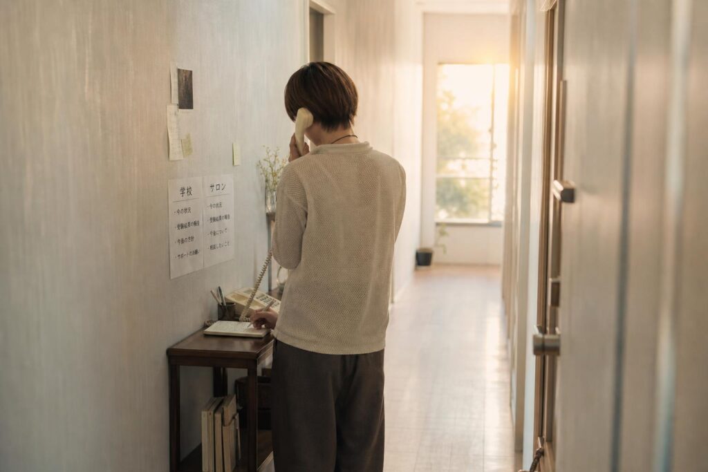 自宅の廊下に置かれた小さな電話台の前で、一人の人物が受話器を耳に当てながら、もう一方の手でメモ帳に「学校」「サロン」と二つの欄を作り、それぞれの下に伝えるべき内容を書き込んでいる。カメラは廊下の奥からの引き気味のロングショットで、電話台に向かう人物の後ろ姿と壁に貼られたメモを同一フレームに収め、背景の窓から差し込む夕方の光が廊下を柔らかく照らす様子がぼんやりと写り込み、二つの相談先に同じタイミングで丁寧に連絡しようとする公平で落ち着いた姿勢を強調する構図になっている。