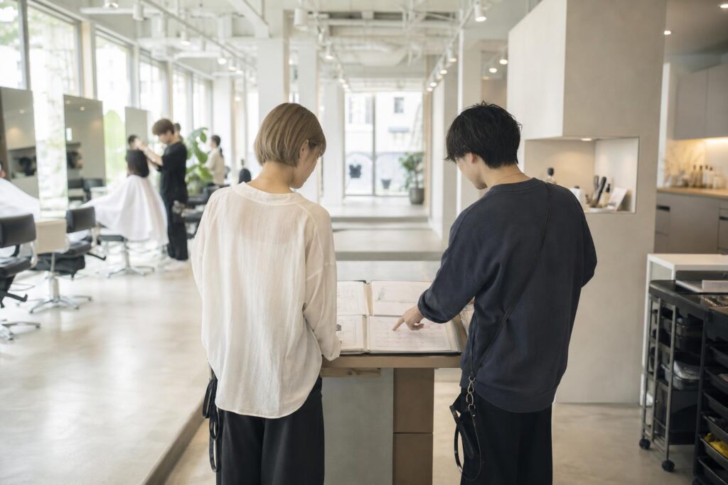 サロンのセット面の脇にある小さな作業台で、退職者役と後任スタイリスト役の二人が並んで立ち、開いた顧客カルテのファイルと予約カレンダーを台の上に広げ、後任側が指先で今後の予約日を一つずつ確認している。カメラは店内奥からの引き気味のロングショットで、二人の後ろ姿と台の上に広がる資料を縦の奥行きの中に収め、背景では別のセット面で施術中のスタッフの気配がぼんやりと写り込み、抱えていた予約を丁寧に次の担当へ渡していく実務的な引き継ぎの様子を強調する構図になっている。