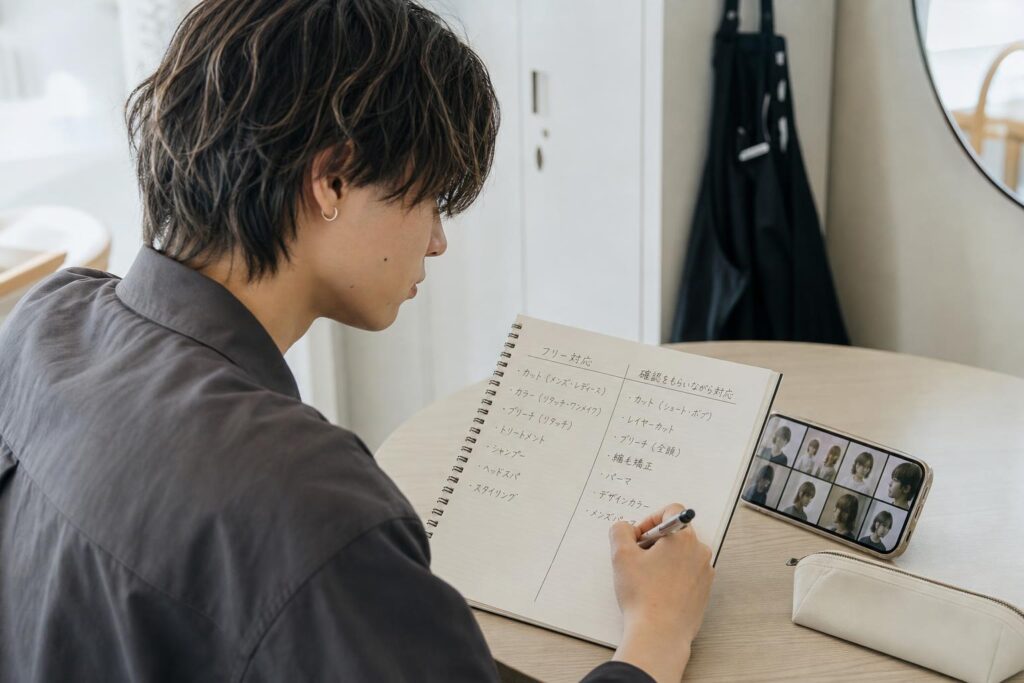 サロンの休憩室にある小さなテーブルで、一人の人物がノートを開き、担当メニューの欄を「フリー対応」と「確認をもらいながら対応」の二列に分けて書き込みながら、隣に置いたスマートフォンで過去の施術写真を見返している。カメラは斜め後方からの近距離バストショットで、ノートの二列の書き分けと人物の横顔を同一フレームに収め、背景にはロッカーに掛けられたエプロンと壁の鏡の端がぼんやりと写り込み、経験の幅と正確さを丁寧に言葉へ落とし込もうとする集中した姿勢を強調する構図になっている。