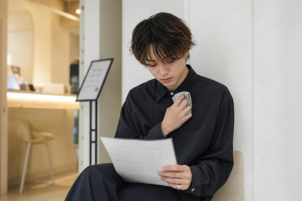 面接会場の外の廊下で、一人の人物が壁際の椅子に浅く腰掛け、膝の上に置いた履歴書のコピーを見つめながら、もう一方の手で胸ポケットのハンカチを握りしめている。カメラは正面やや低めからのクローズアップショットで、俯いた表情と履歴書の文字がぼやける様子を同一フレームに収め、背景には受付カウンターの淡い照明と順番待ちの案内板がぼんやりと写り込み、面接直前の緊張と拭えない不安が入り混じる様子を強調する構図になっている。