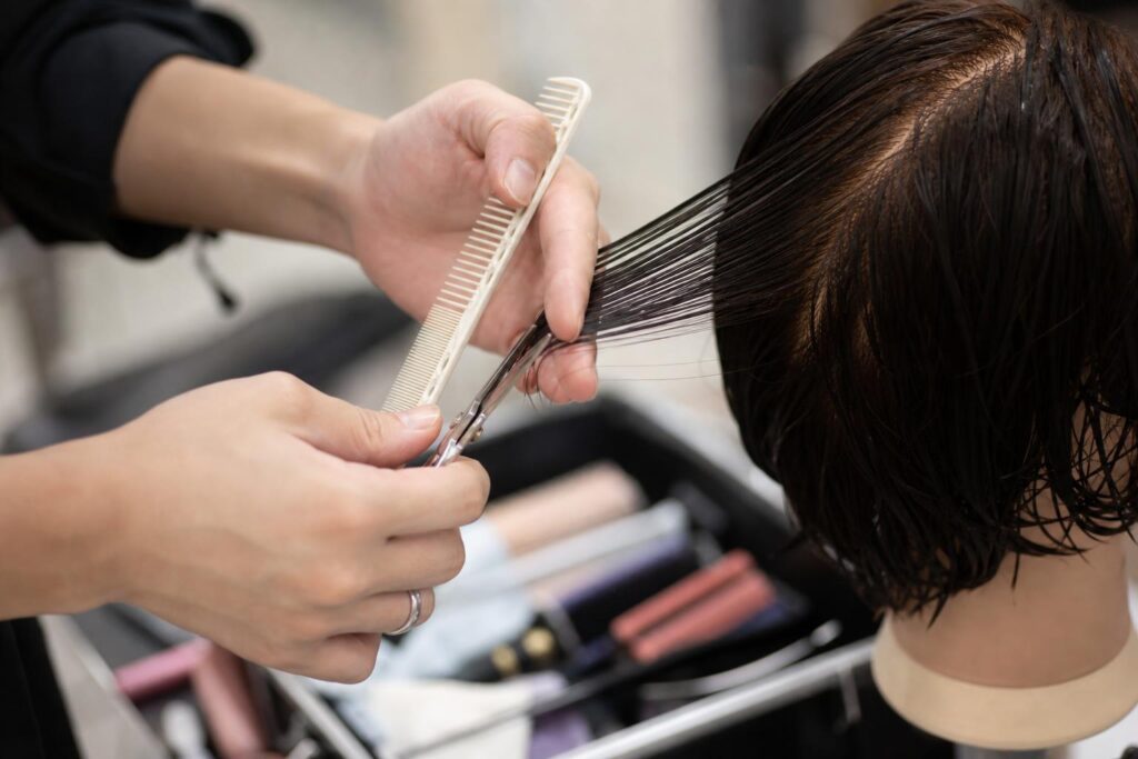 美容室の作業台で、美容師役の人物が撮影用のウィッグに向かいハサミとコームを使い技術チェックの練習をしている。カメラは手元へ寄った近距離ショットで、顔は入れず指先と刃先の動きだけを鮮明に捉え、背景の道具箱がわずかにぼける構図。