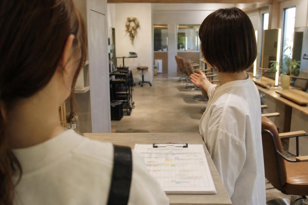 サロン見学に訪れた人物の肩越しの視点で、受付奥に広がる予約表とセット面を眺める場面を捉える。案内するスタッフの後ろ姿と客席の配置を中距離で写し、紙面の文字は判読できないほどぼかした構図。