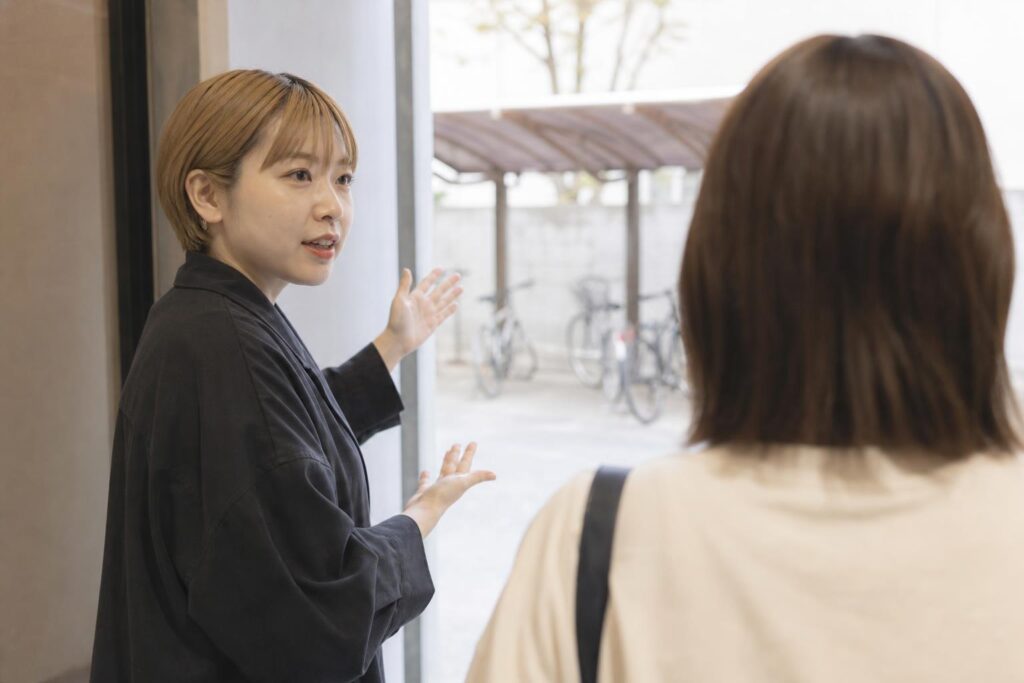 サロンの裏口手前で、応募者役の肩越しに案内役スタッフがジェスチャーを交えてSNS運用の担当範囲を説明する様子を中距離で捉え、背景の自転車置き場がぼんやり写り込む構図。