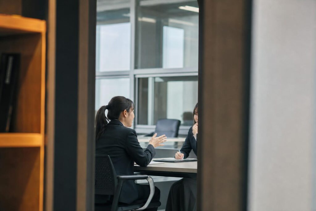 面接ブースの入口枠を前景に使い、応募者役が向かいの人事担当者へ身を乗り出して質問を投げかける様子を画面中央から外れた位置で中距離から捉え、背景の窓枠と椅子の輪郭がぼんやり写り込む構図。