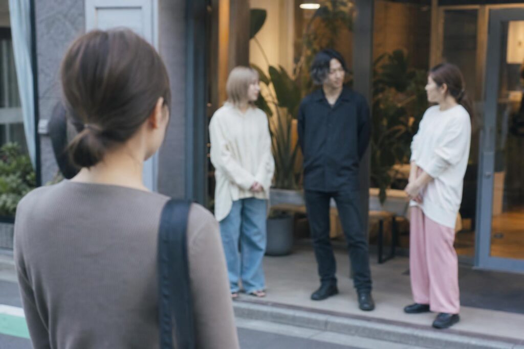 商店街の歩道で、応募者役の肩越しに、サロン入口に立つスタッフたちの私服姿を中距離で眺める様子を捉え、背景のガラス扉と観葉植物の輪郭を柔らかくぼかして写す構図。