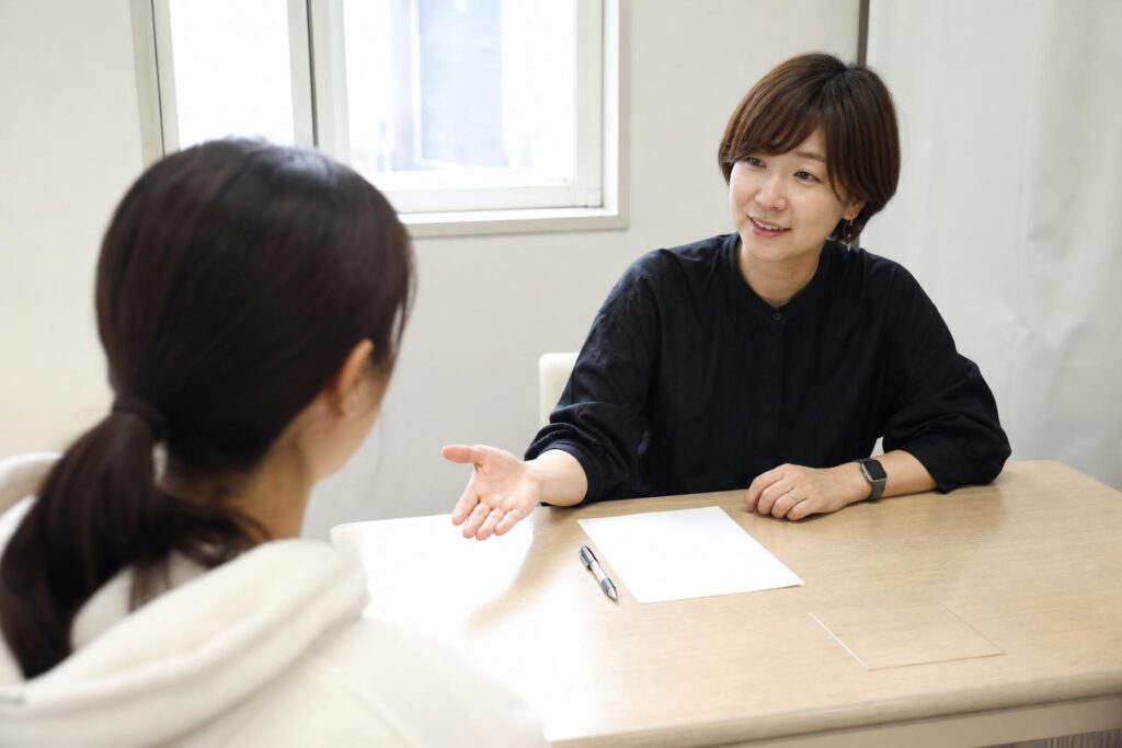 美容学校の進路相談室で、学生役を画面左手前に後ろ姿で置き、その肩越しに、机の向かいで先生役が片手を開いて穏やかに助言する様子をやや高い位置から中距離で捉える。机上は無地の紙とペンのみ、窓の光で文字は見せない。