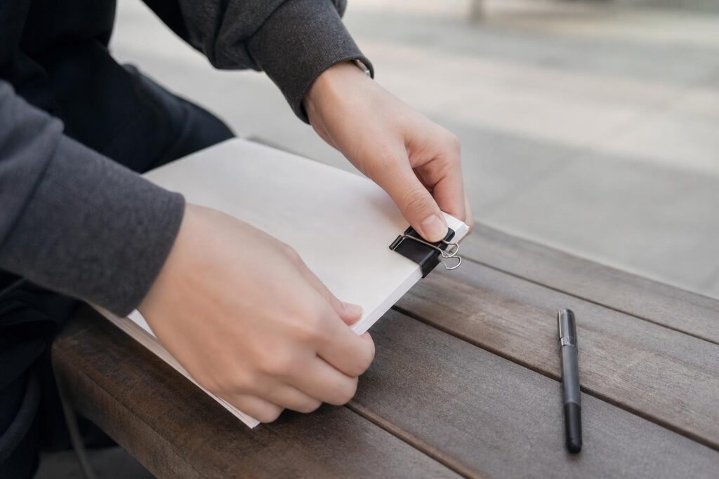 会場脇のベンチ上を、座面の高さの斜め上から寄りで撮る。顔は入れず、裏向きに重ねた無地の冊子の端を片手で揃え、もう片手が黒いクリップで留める。脇にペン一本。背景は淡くぼかし、文字は見せない。