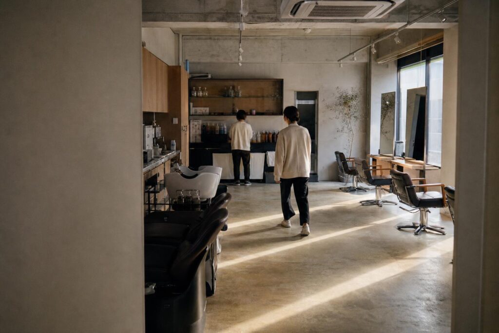 閉店後のサロンを、シャンプー台側の入口から広く引いて撮る。画面右三分の一でスタッフ役が小さく歩み出し、奥の棚でカラー剤のボトルを揃える店長役の背へ近づく。窓の斜光が床に伸びて奥行きが出る。文字は写さない。