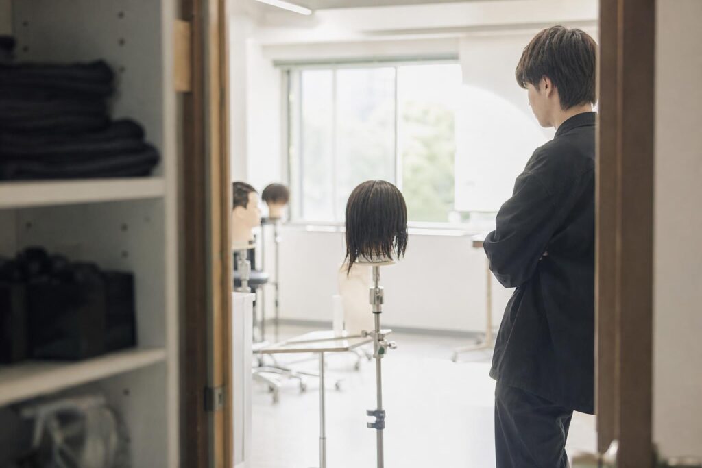 朝の美容学校の実習室を、廊下側のドア枠越しに目線の高さで撮る。枠の右端に学生役が横向きで立ち、マネキンから一歩下がって全体の左右バランスを静かに見る。中央は窓の朝光で空け、文字は写さない。