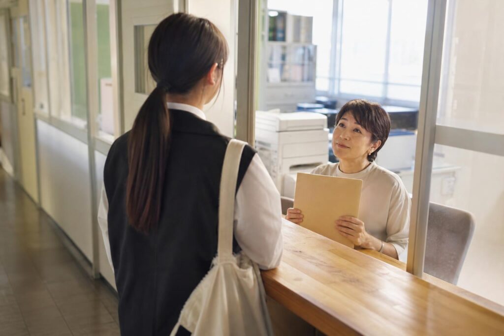 放課後の学校の職員室前の木製カウンターで、学生役を画面中央やや左に後ろ姿で置く。肩越しに、椅子の先生役が顔を上げ、閉じた無地のファイルを両手で持ち穏やかに頷く。やや高い位置から中距離で、窓の淡い夕光、文字なし。