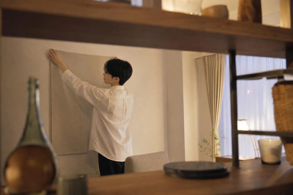 夕方の自宅の一室を、手前の木製棚の段の隙間越しに目線の高さで撮る。画面左寄りで白シャツの応募者役が横向きに立ち、背後の無地の壁に掛けた布を真っすぐに直す。窓の淡い光、文字なし。