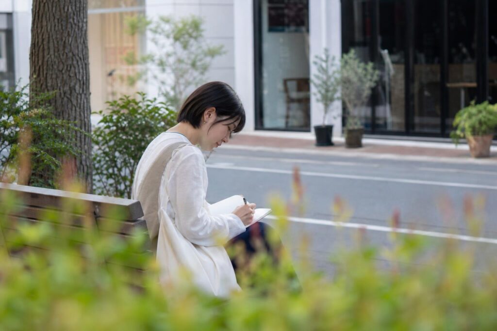 昼下がり、サロンのある通りの向かい側から、低い花壇の葉を前景に淡くぼかし望遠で撮る。街路樹の下のベンチで、見学帰りの学生役が横向きに座り、膝の無地の手帳へ静かに書き留める。文字なし。