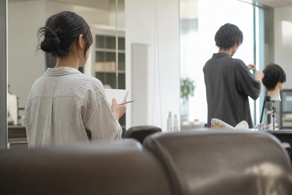 昼のサロンのフロアを、客席の背もたれを前景に淡くぼかし、望遠で斜め後ろから静かに撮る。中央やや左で学生役が立ち、小さな無地のノートに鉛筆を添える。奥で離れて美容師役がマネキンの髪を整える。文字なし。